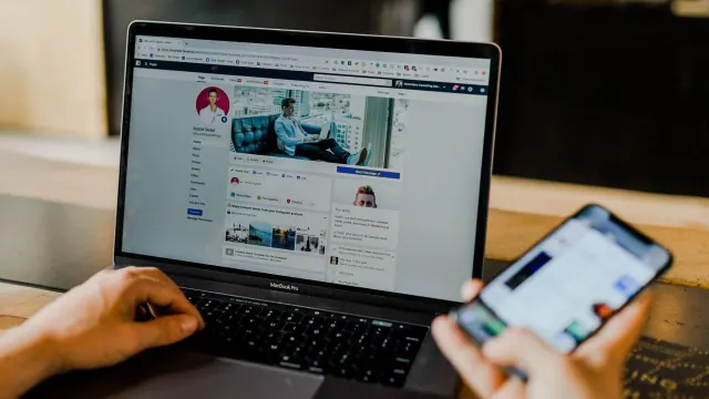 A person looking at their laptop and phone. The laptop has Facebook open on someone’s profile page. The phone screen is blurred.
