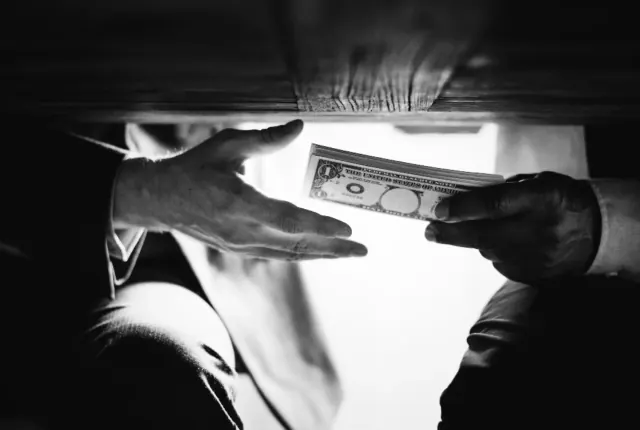 A man is pictured holding numerous American bills to another under a table