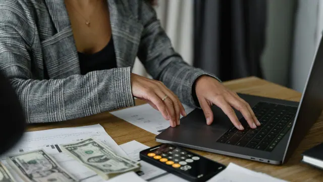 A person in a gray plaid blazer is typing on a laptop. On the wooden desk, there are scattered U.S. dollar bills, a calculator, and printed financial graphs, suggesting a financial planning or budgeting activity