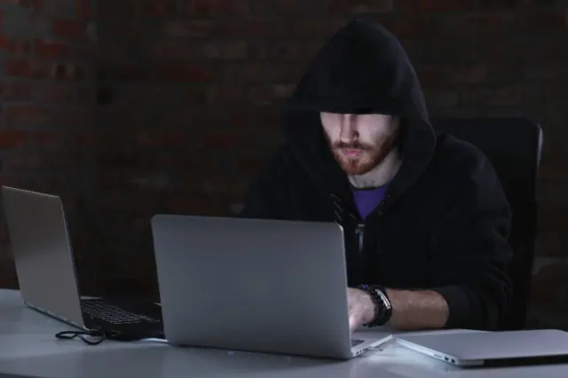 A man in a black hoodie is pictured using his laptop for illicit purposes