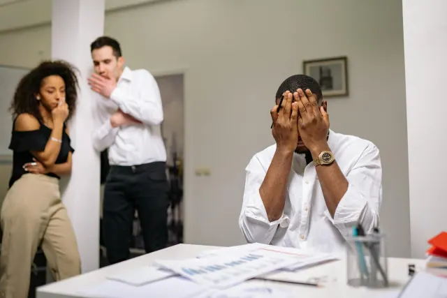 A man in a white shirt sitting covering his face while another man and a woman standing behind him beside a pillar