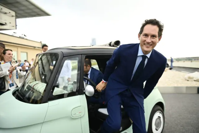 A man in a suit smiles beside the open door of a small car.