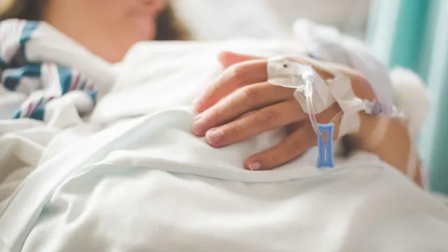 A person lying in a hospital bed with an IV attached to their hand.