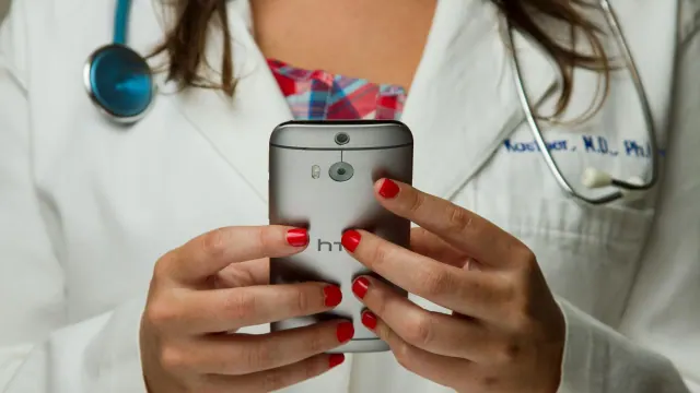 A doctor wearing a stethoscope and a white lab coat on her phone.