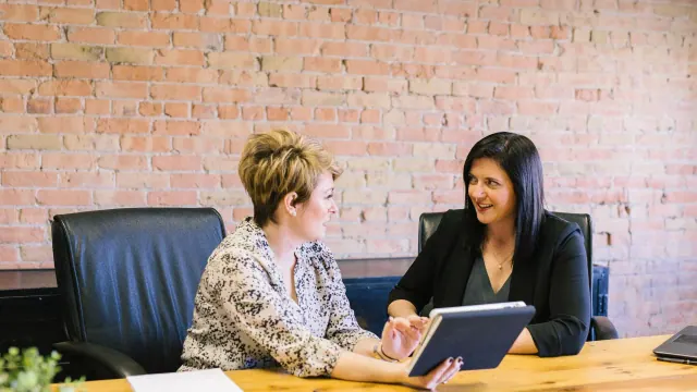Two people discussing paperwork and a tablet at a desk