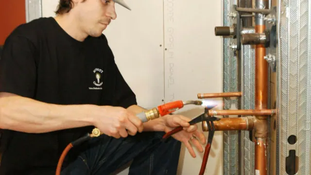 A worker holding a torch beside exposed copper pipes