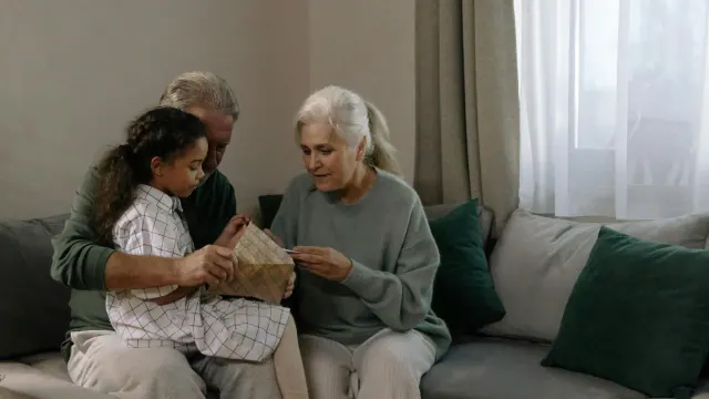 A photo showing an elderly couple with a young girl, presumably a granddaughter. The girl is sat on the knee of the man and they are all engaged in opening a card