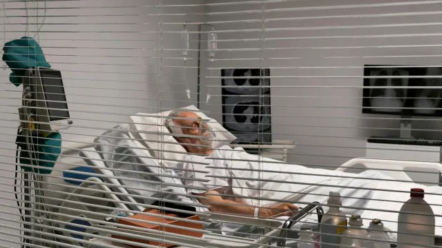 View through window blinds of a hospital room, where an elderly patient is lying in bed with a head covering and a mask, next to medical equipment