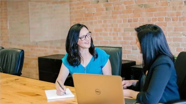 Two women sit at a table with a laptop and a pen and pad of paper, a brick wall behind them.