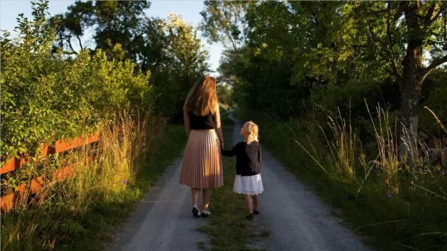 A mother and daughter walking down a path surrounded by green trees and bushes.