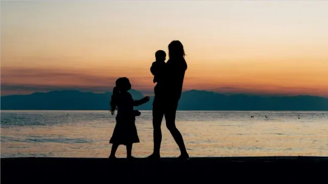 Silhouettes of a mother with her two children, one she is holding, in front of a body of water during a sunset.