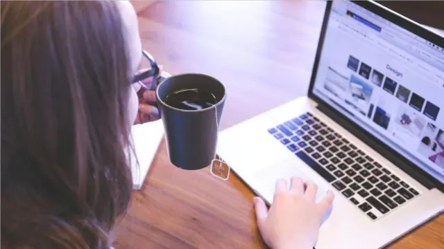 A woman in glasses uses a laptop while holding a mug full of tea.
