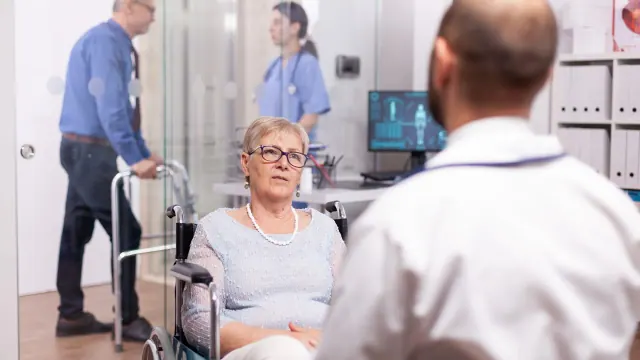 An elderly female patient in a wheelchair engaged in a conversation with a doctor in a medical facility. In the background, a male nurse is seen pushing a walker