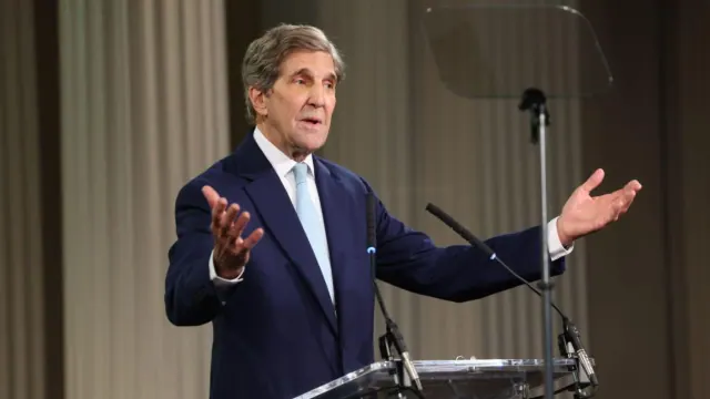 John Kerry speaking at a microphone with both hands raised
