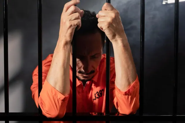 A man pictured in an orange jumpsuit holds on to the bars of his prison cell