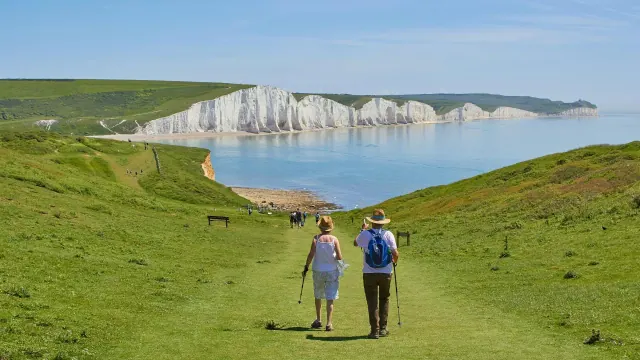 An elderly couple holding walking sticks are seen from behind, strolling down a grassy path towards a beach