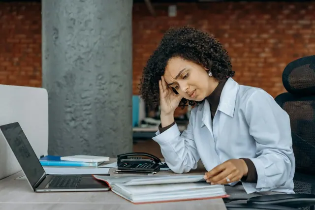 A curly-haired woman with a serious expression reading from a file with a laptop open on the table in front of her