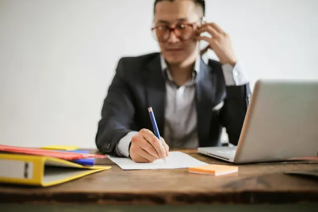 A man wearing glasses sitting at a table speaking on the phone while writing on paper