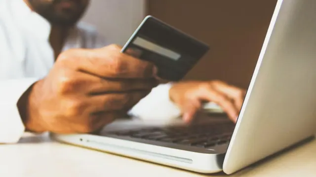 A man sat at his desk with a laptop and credit card. He is holding the card and is putting the numbers from it into his laptop.