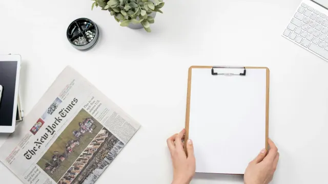 A person holds a blank clipboard at a desk beside a newspaper and plant.