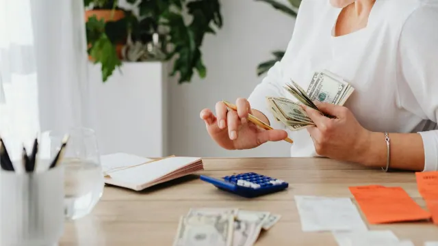A woman is holding a stack of US currency and a pen. She is actively using a calculator and has a notebook and receipts on the desk in front of her