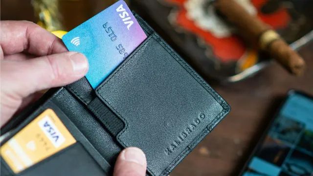 A person's hand is pulling out a blue Visa credit card from a black leather wallet