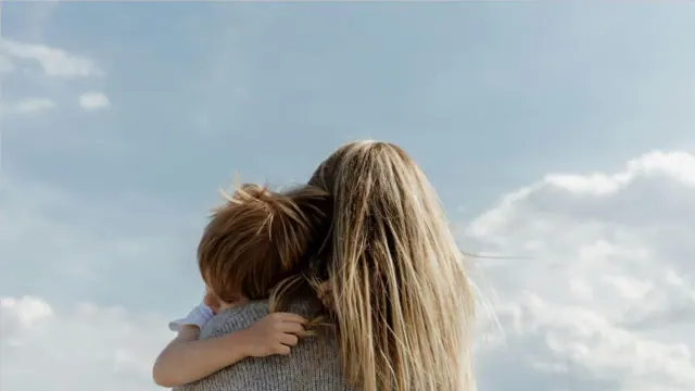 A mother holds her child outside in the daytime under a blue and cloudy sky.