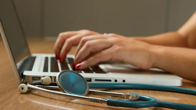 A person typing on a laptop on a wooden surface with a stethoscope on the table next to them.