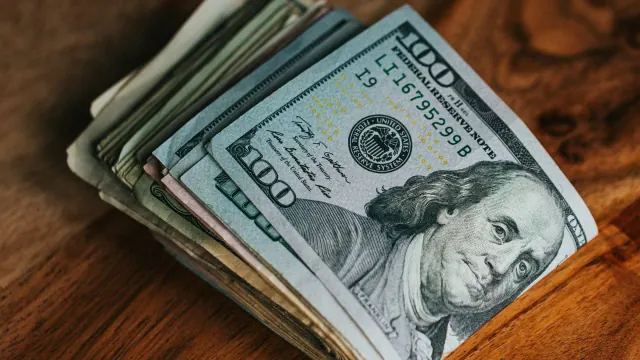 A close-up image showing a stack of United States currency, specifically hundred dollar bills, with the face of Benjamin Franklin visible, placed on a wooden surface