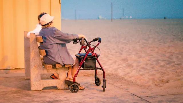 A person sitting on a bench beside a wheeled walking aid near a beach