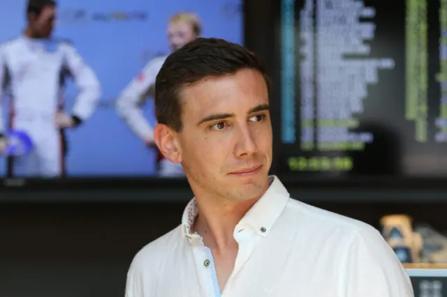 A man in a white shirt looks to one side in front of a blurred display.