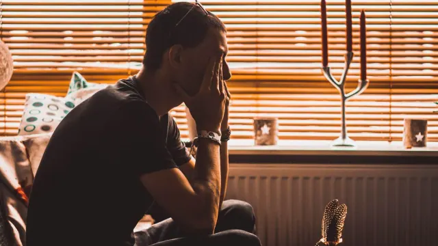 A man with his head in his hands. He has the blinds shut and is on his own