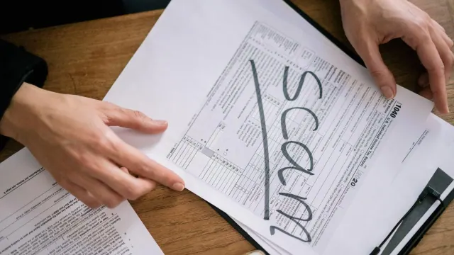 A person looking through some paper documents. The piece of paper on the top has ‘scam’ written in grey writing and has been underlined.